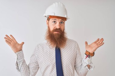 İzole edilmiş beyaz arka plan üzerinde güvenlik kaskı takan genç kızıl saçlı mimar elleri ve kolları havada, kafası karışmış bir ifade takınıyor. Şüphe kavramı.