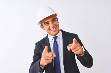 İzole edilmiş beyaz arka plan üzerinde kask ve takım elbiseli genç, yakışıklı mimar mutlu ve komik yüzlü parmaklarını kameraya doğrultuyor. İyi enerji ve titreşimler.