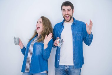 Genç ve güzel bir çift beyaz izole arka planda bir fincan kahve içerken mutlu ve heyecanlı, kazanan yüz ifadesi zaferi büyük bir gülümsemeyle kutluyor ve ellerini kaldırıyor.