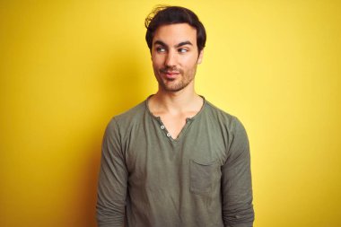 Genç yakışıklı bir adam günlük tişört giyip izole edilmiş sarı arka planda duruyor gülümsüyor ve başka tarafa bakıyor..