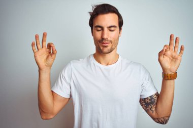 İzole edilmiş beyaz arka plan üzerine gündelik tişört giyen yakışıklı genç adam rahatla ve gözün kapalı gülümse parmaklarınla meditasyon hareketi yap. Yoga konsepti.