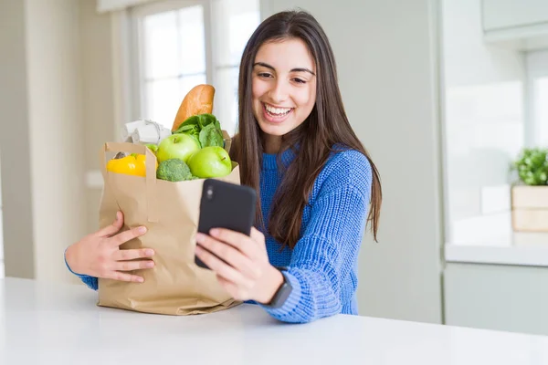Young woman holding a paper bag full of fresh groceries and using smartphone app for supermarket delivery