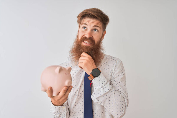 Young redhead irish businessman holding piggy bank over isolated white background serious face thinking about question, very confused idea