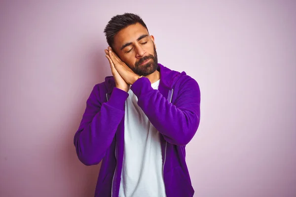 Young indian man wearing purple sweatshirt standing over isolated pink background sleeping tired dreaming and posing with hands together while smiling with closed eyes.