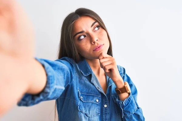 Beautiful woman wearing denim shirt make selfie by camera over isolated white background with hand on chin thinking about question, pensive expression. Smiling with thoughtful face. Doubt concept.