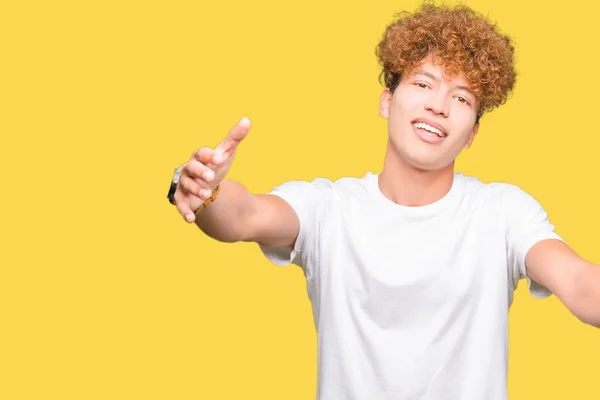 Young handsome man with afro hair wearing casual white t-shirt looking at the camera smiling with open arms for hug. Cheerful expression embracing happiness.