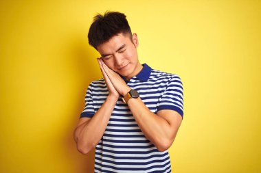 Çizgili polo giyen Asyalı genç adam izole edilmiş sarı arka planda duruyor. Yorgun uyuyor, hayal kuruyor ve ellerimiz kapalı gülümserken birlikte poz veriyor..