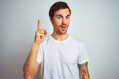 Başarılı bir fikir ile parmak işaret izole beyaz arka plan üzerinde rahat t-shirt giyen dövme ile genç yakışıklı adam. Çıktı ve mutlu. Bir numara.
