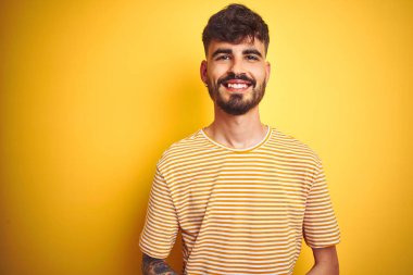 İzole edilmiş sarı arka plan üzerinde duran çizgili tişörtlü genç adam ellerini çapraz yapıp gevşemiş ve neşeli gülümsüyor. Başarı ve iyimser