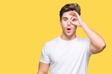 Sürpriz yüz, göz parmaklarının arasından seyir ile ok jest yapıyor izole arka plan üzerinde beyaz t-shirt giyen genç yakışıklı adam şok. İmansız ifade.