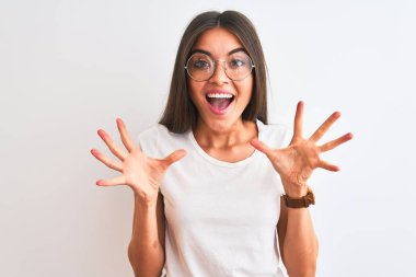 Genç ve güzel bir kadın günlük tişört ve gözlük takıp izole edilmiş beyaz arka planda çılgınlığı kutluyor ve başarıya hayret ediyor. Kolları havada ve gözleri açık, heyecanlı bir şekilde çığlık atıyor. Kazanan konsept