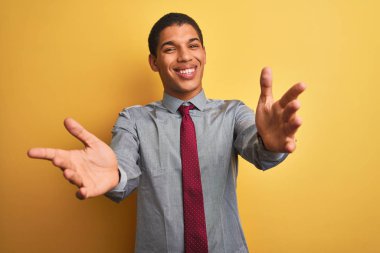 Yakışıklı Arap iş adamı gömleği ve kravat takıp izole edilmiş sarı arka planda kameraya sarılıp gülümsüyor. Mutluluğu kucaklayan neşeli bir ifade..