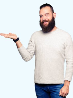 Neşeli sunulması ve kameraya bakarak avucunun ile işaret gülümseyen rahat kazak giyen genç sarışın adam.