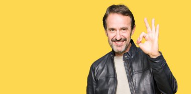 Orta yaş yakışıklı adam el ve parmaklar ok işaretiyle yapıyor olumlu gülümseyen siyah deri ceket giyiyor. Başarılı ifade.