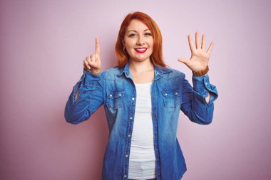 Genç, güzel, kızıl saçlı, kot gömlek giyen, pembe arka planda duran, 6 numaralı parmaklarıyla işaret eden, kendinden emin ve mutlu bir şekilde gülümseyen kadın..