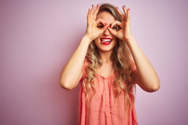 Pembe izole edilmiş arka planda duran tişört giyen genç ve güzel bir kadın dürbünün dili dışarı çıkarması, gözlerin parmaklarından içeri bakması gibi güzel bir jest yapıyor. Çılgın ifade.