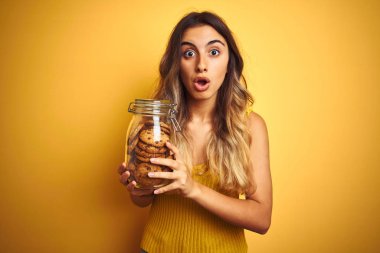 Sarı arka planda bir kavanoz kurabiyeyle gezen genç ve güzel bir kadın. Şaşırmış bir yüzle korkmuş ve korku ifadesinden heyecanlanmış.