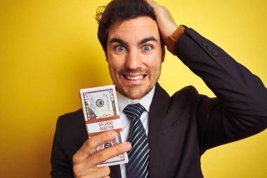 Takım elbiseli genç yakışıklı işadamı, dolar tutarak izole edilmiş sarı arka planda, elleri başının üstünde, utanç ve şaşkınlık dolu, sinirli ve hayal kırıklığına uğramış bir halde. Hata için korku ve üzüntü.