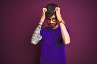 Dövmeli, tişörtlü ve gözlüklü genç bir adam izole edilmiş mor arka planda duruyor. Ağrı ve migren yüzünden baş ağrısı ve stres altında. Ellerini başının üstüne koy..