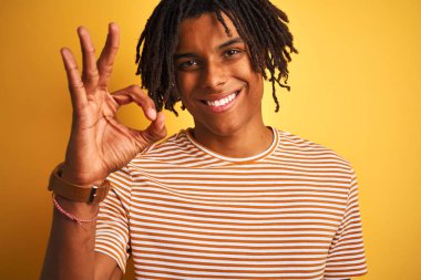 Rastalı tişörtlü Afro adam izole edilmiş sarı arka planda duruyor. Parmaklarıyla imza atıyor. Mükemmel bir sembol.