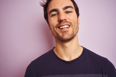 İzole edilmiş pembe arka plan üzerinde duran sıradan tişört giyen genç yakışıklı bir adam mutlu bir yüzle ayakta duruyor ve gülümsüyor dişlerini gösteren bir gülümsemeyle.