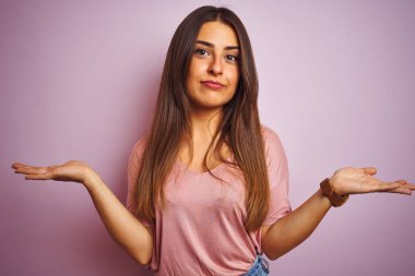 İzole edilmiş pembe arka planda duran tişört giyen genç ve güzel bir kadın. Kolları ve elleri havada, kafası karışmış bir ifade. Şüphe kavramı.