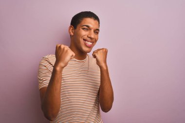 İzole edilmiş pembe arka plan üzerinde duran çizgili tişörtlü genç yakışıklı Arap adam kollarını kaldırarak, gülümseyerek ve başarı için bağırarak mutlu ve heyecanlı bir jest yapıyor. Kutlama konsepti.