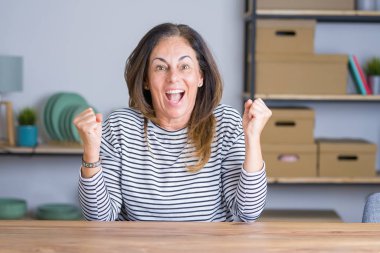 Evde masada oturan orta yaş kıdemli kadın şaşırttı ve silah kaldırdı ve açık gözleri ile başarı için hayran kutluyor. Kazanan kavramı.
