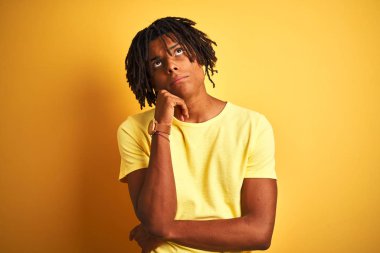Rastgele tişörtler giyen Afro bir adam izole edilmiş sarı arka planda duruyor, elleri çenesinde soru sormayı düşünüyor, düşünceli bir ifade. Düşünceli bir yüzle gülümsüyor. Şüphe kavramı.