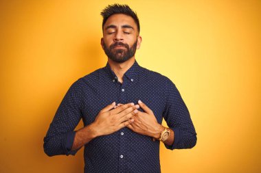 Genç, yakışıklı Hintli iş adamı, izole edilmiş sarı arka planda tişört giyiyor, elleri göğsünde, gözleri kapalı ve yüzünde minnet dolu bir jest ile gülümsüyor. Sağlık kavramı.