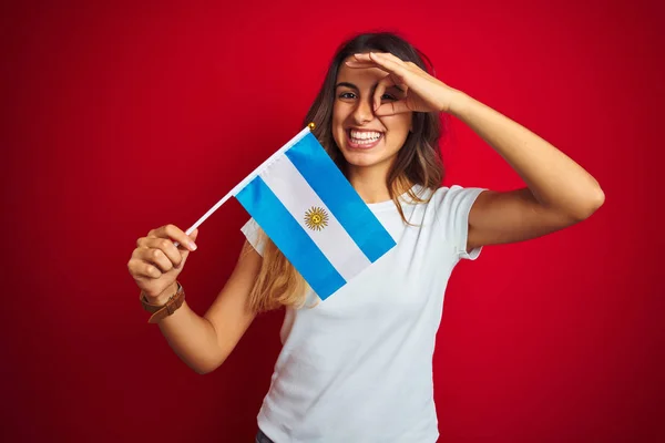 若いです美しいです女性保持Argentineフラグ上の赤隔離された背景とともに幸せな顔笑顔行いますOk Signとともに手で目を通して指を見て — ストック写真