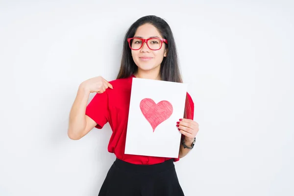 Young brunette woman holding card with red heart over isolated background with surprise face pointing finger to himself