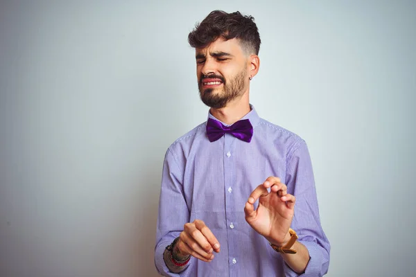 Young man with tattoo wearing purple shirt and bow tie over isolated white background disgusted expression, displeased and fearful doing disgust face because aversion reaction. With hands raised. Annoying concept.