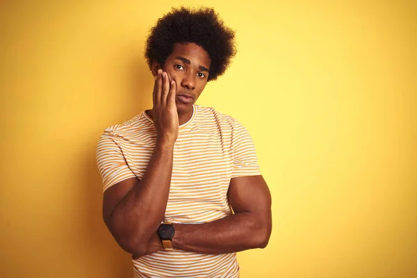 American man with afro hair wearing striped t-shirt standing over isolated yellow background thinking looking tired and bored with depression problems with crossed arms.