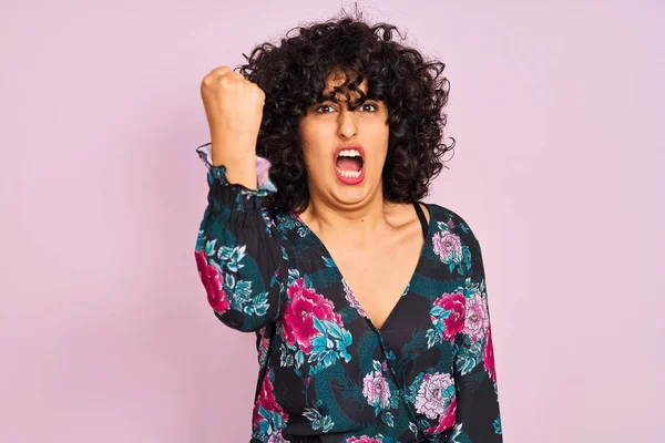 Young arab woman with curly hair wearing floral dress over isolated pink background angry and mad raising fist frustrated and furious while shouting with anger. Rage and aggressive concept.