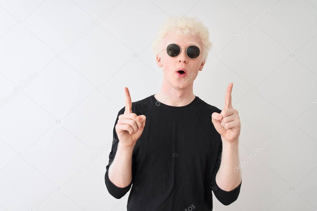 Young albino man wearing black t-shirt and sunglasess standing over isolated white background amazed and surprised looking up and pointing with fingers and raised arms.