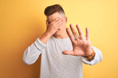 Beyaz tişört giyen yakışıklı genç adam izole edilmiş sarı arka plan üzerinde duruyor gözleri ellerle örtüyor ve üzgün ve korku ifadeleriyle jest yapıyor. Utanç verici ve olumsuz bir kavram..