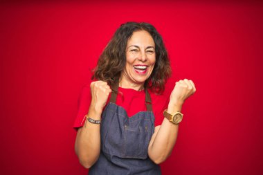 Kırmızı izole edilmiş arka planda önlük giyen orta yaşlı bir kadın mutlu ve heyecanlı bir şekilde kollarını kaldırıp, gülümseyerek ve başarı için çığlık atarak zafer jesti yapıyor. Kutlama konsepti.