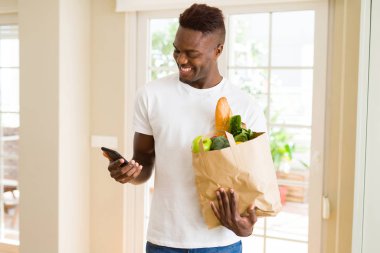Bakkaliye tam bir kağıt torba tutarak ve online satın alma smarpthone kullanarak Afrika adam gülümseyerek app kullanarak