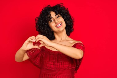 Kıvırcık saçlı genç bir Arap kadın izole edilmiş kırmızı arka planda gündelik bir elbise giyiyor. Aşkla gülümsüyor. Elleriyle kalp sembolü ve şekli gösteriyor. Romantik konsept.