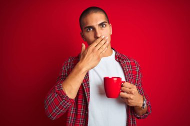 Gömlek giyen genç yakışıklı bir adam kırmızı izole edilmiş arka planda bir fincan kahve içiyor. Ağzı kapalı. Yanlışlıkla utancından ağzı sarsılıyor.