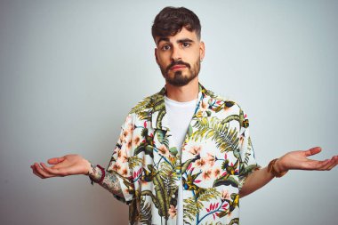 Tatilde dövmeli bir adam, izole edilmiş beyaz arka plan üzerinde duran kolları ve elleri havada, kafası karışmış bir adam. Şüphe kavramı.