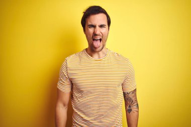 İzole edilmiş sarı arka plan üzerine çizgili tişört giyen yakışıklı genç adam mutlu bir ifadeyle dilini dışarı çıkarıyor. Duygu kavramı.