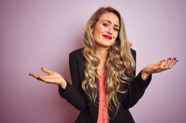 Pembe izole edilmiş arka plan üzerinde duran zarif bir ceket giyen genç ve güzel bir iş kadını. Kolları ve elleri havada, kafası karışmış bir ifade. Şüphe kavramı.
