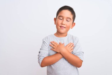 Gri günlük tişört giyen güzel çocuk izole edilmiş beyaz arka plan üzerinde duruyor elleri göğsünde, gözleri kapalı ve yüzünde minnet dolu bir jest ile gülümsüyor. Sağlık kavramı.