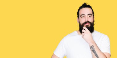 Uzun saç ve sakal emin çapraz kollar ve çeneme kaldırdı el ile gülümseme ile kameraya bakarak casual beyaz t-shirt giyen genç hipster adam. Pozitif düşünce.