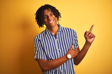 Rastgele çizgili tişört giyen Afro bir adam izole edilmiş sarı arka planda, yüzünde kocaman bir gülümseme, eli ve parmağı kameraya bakıyor..