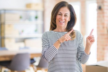 Güzel orta yaşlı bir kadın ayakta gülümsüyor ve kameraya bakıyor iki eliyle ve parmaklarıyla yan tarafa bakıyor..