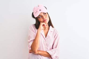 İzole edilmiş beyaz arka plan üzerinde uyku maskesi ve pijama giyen genç ve güzel bir kadın. Elleri çenesinde soru sorarken, düşünceli bir ifade takınıyor. Düşünceli bir yüzle gülümsüyor. Şüphe kavramı.
