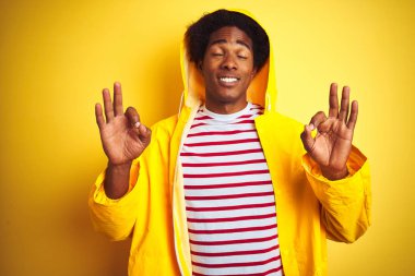 Kapüşonlu bir yağmurluk giyen afro saçlı Afrikalı adam izole edilmiş sarı arka planda duruyor rahatla ve gözleri kapalı meditasyon hareketi yaparken parmaklarınla gülümse. Yoga konsepti.